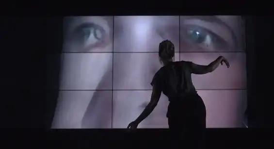 Work-in-progress preview of SELF ABSORBED by Tim Murray-Browne. A woman stands in front of a large video wall striking a pose with her arms. On the video wall is a 3D render of a distorted male face.