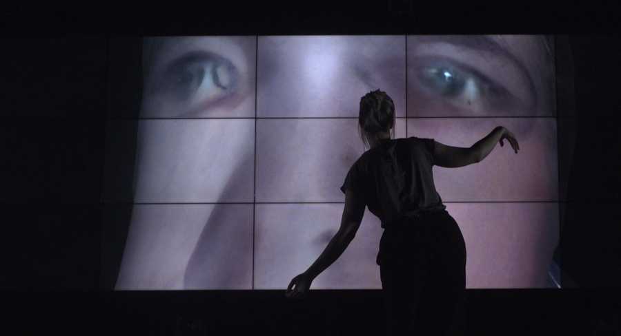 Work-in-progress preview of SELF ABSORBED by Tim Murray-Browne. A woman stands in front of a large video wall striking a pose with her arms. On the video wall is a 3D render of a distorted male face.