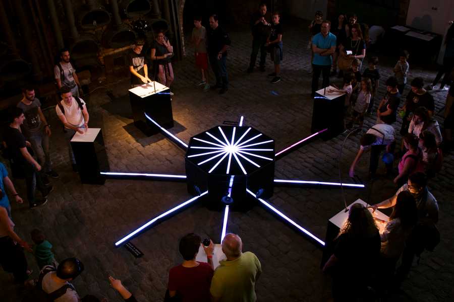 Top down photo of the Cave of Sounds by Tim Murray-Browne and Music Hackspace Ensemble with lots of people interacting. Eight novel instruments are arranged in a circle with a glowing octagonal core. Photo by Anastasia Alekseeva.