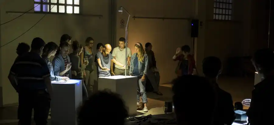 The interactive sound installation “Cave of Sounds” Tim Murray-Browne and the Music Hackspace Ensemble. A man stands in front of a plinth casting shadows on it with his hands, at the edge of an art installation. People stand around watching him. Photo: Tim Murray-Browne