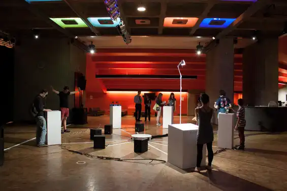 Cave of Sounds exhibited at the Barbican 2013. The photo shows a circle of plinths facing inwards with different Digital Musical Instruments with various people playing them. Photo: Tim Murray-Browne.