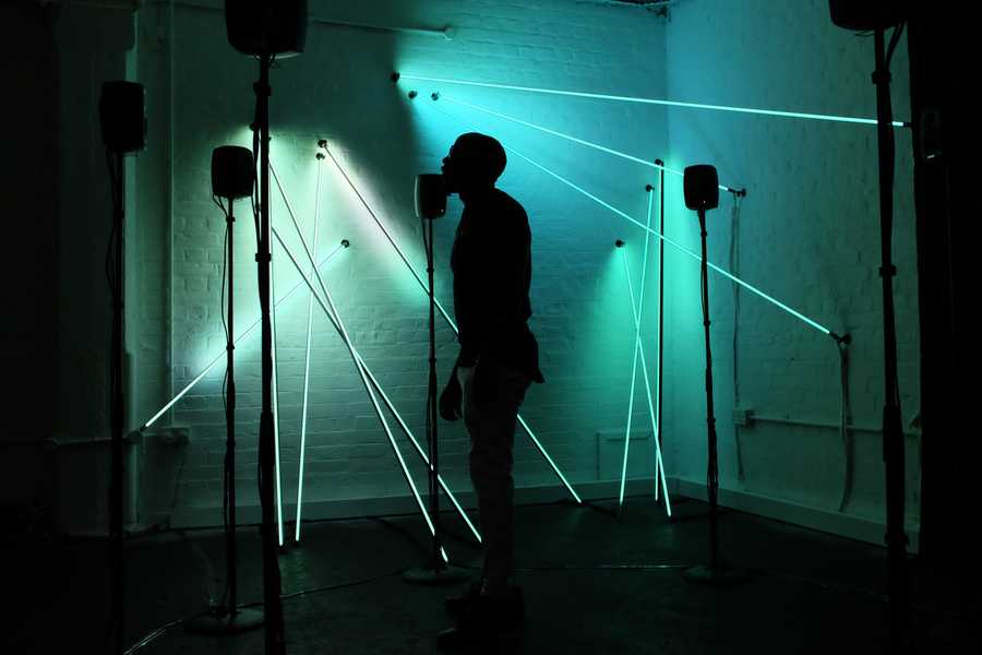 Photo of a man in the ring of speakers in front of luminescent rods that make up the interactive audio-visual sculpture Post Truth and Beauty by Tim Murray-Browne and Aphra Shemza. Photo by Tim Murray-Browne.
