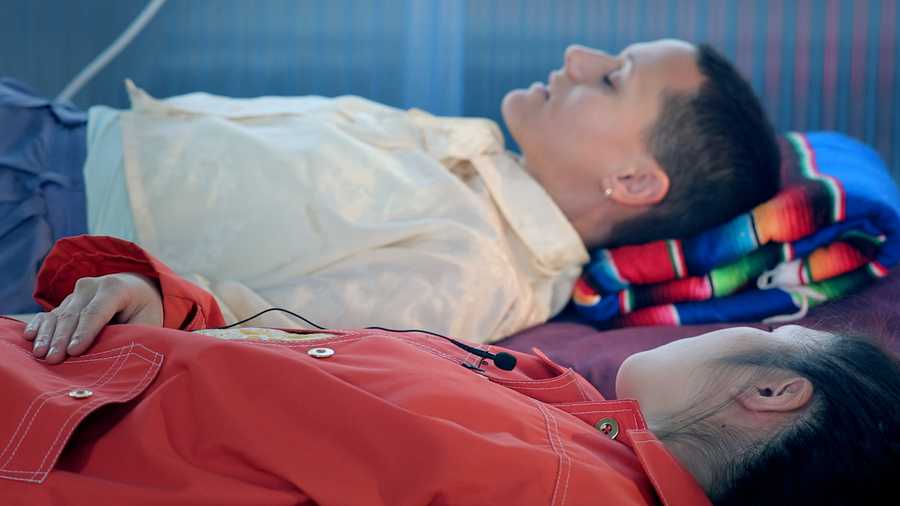 A video still showing the participant lying down with her eyes closed while the host lies next to her. Taken from a film created by Mind The Film of Movement Alphabet by Jan-Ming Lee and Tim Murray-Browne